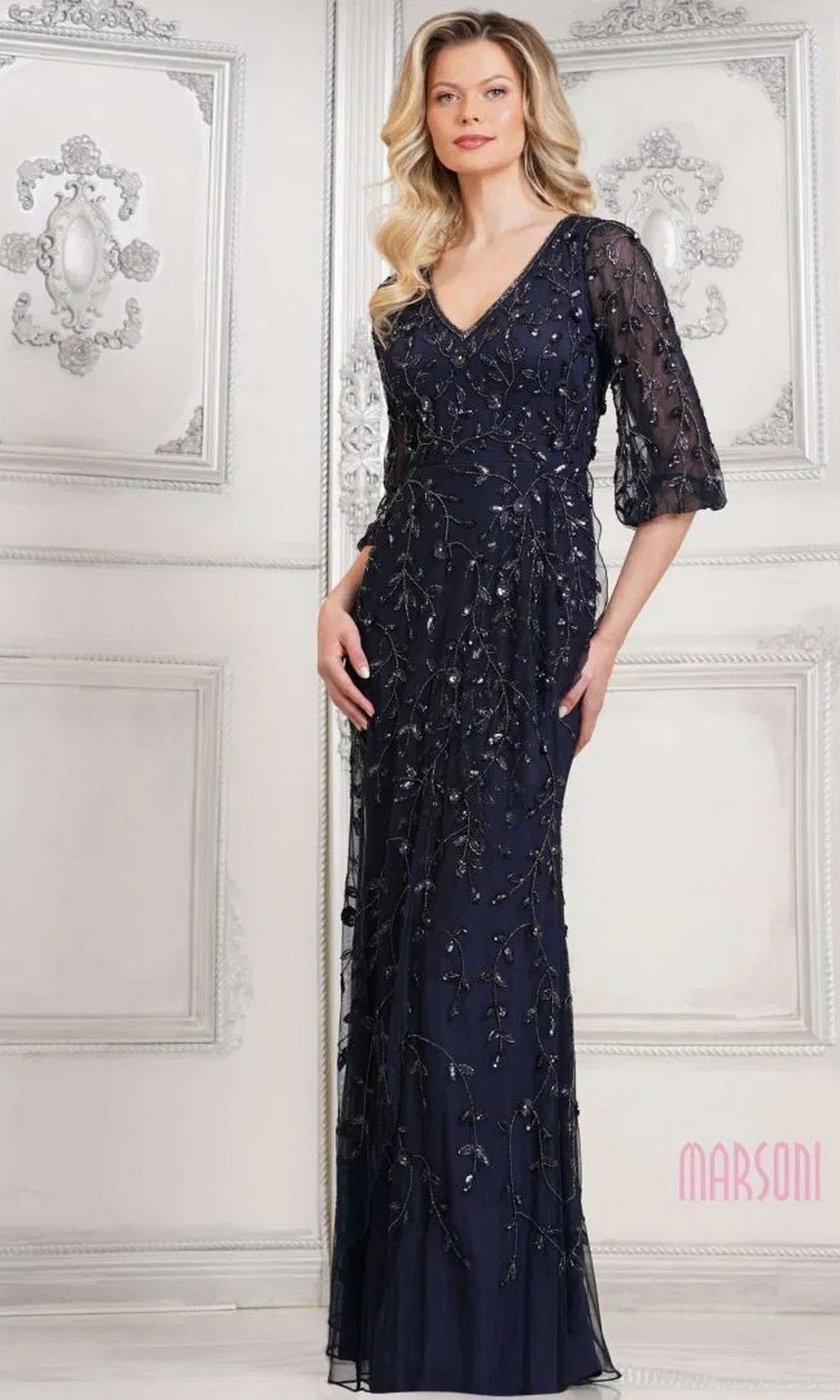 A woman stands in a Marsoni MV1320 long formal navy dress with sequin beading and sheer elbow-length sleeves, set against an ornate white wall.